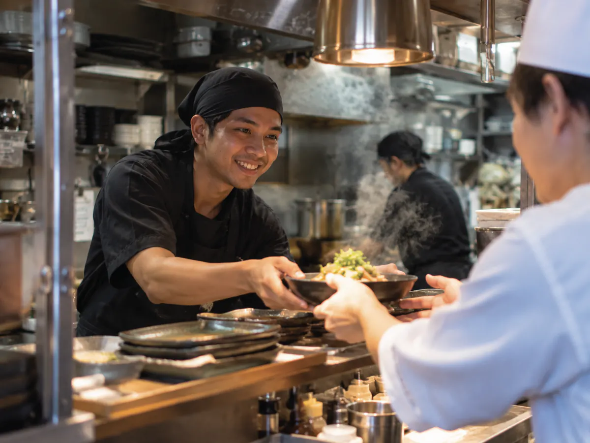 日本料理店の厨房で、外国人の調理スタッフが皿を仕上げ、日本人の同僚が受け取ろうとしている