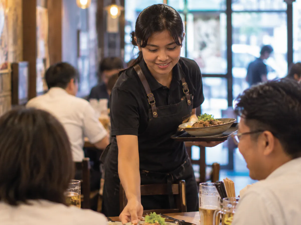 居酒屋のホールで、外国人スタッフが料理を席に運んでいる